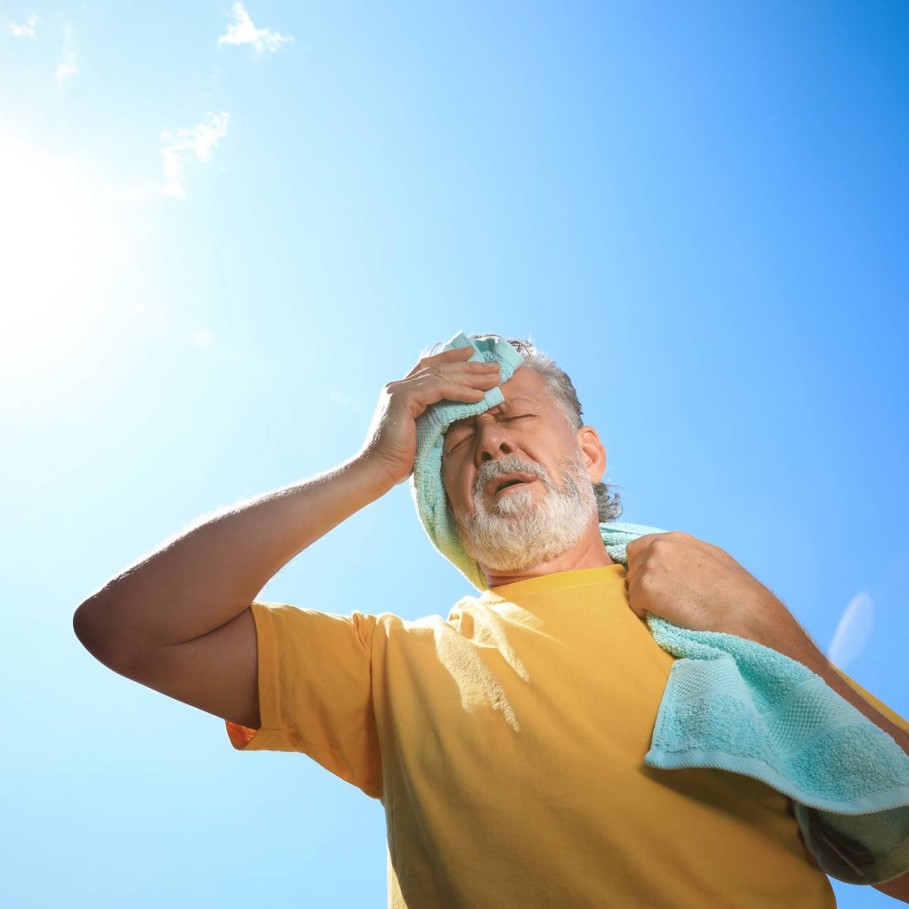 An older man wipes sweat off his brow, carrying a towel outside, bright sun overhead