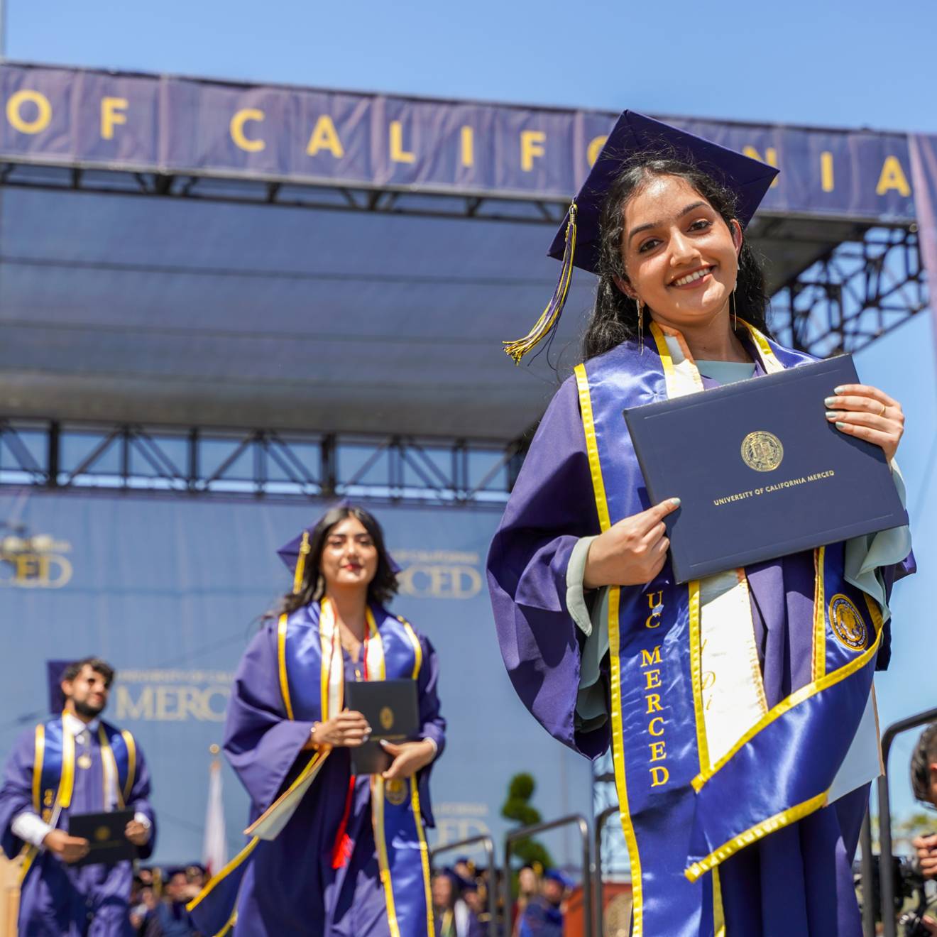 Three UC Merced graduates hold their diplomas as they leave the stage of graduation, smiling