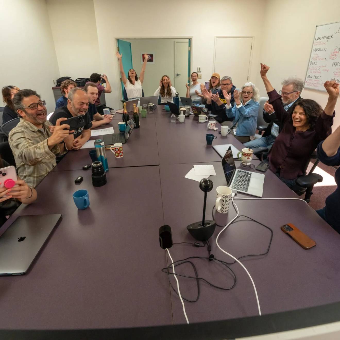 A boardroom table full of people celebrates at Pulitzer Prize win, taking selfies, raising arms, shouting, etc.