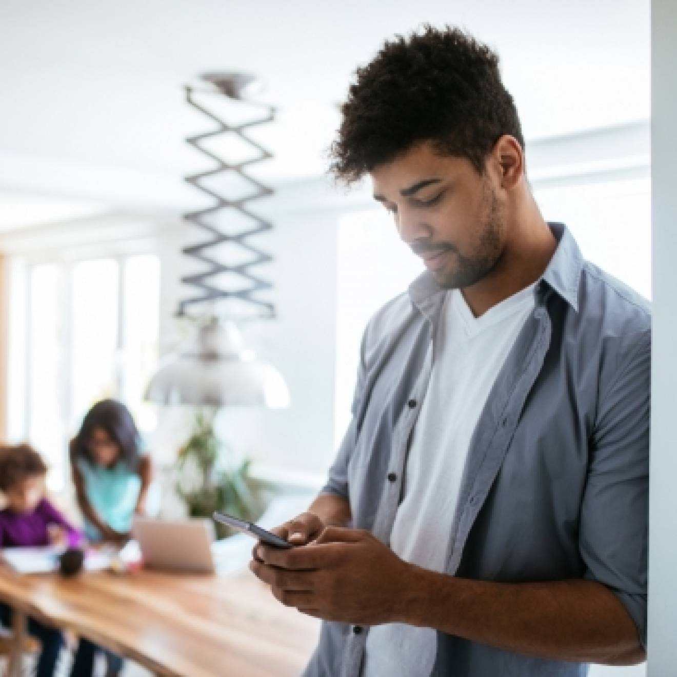 A man uses a smartphone while his kids are at a table in the background