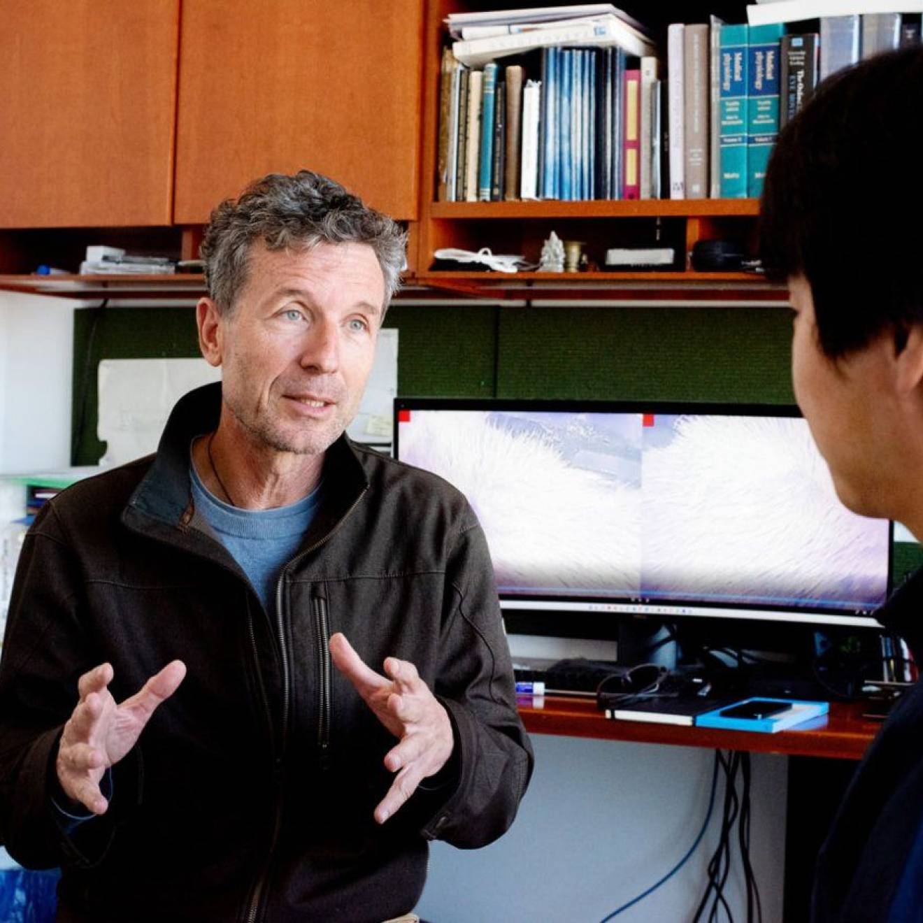A researcher with gray hair at his desk talks to someone