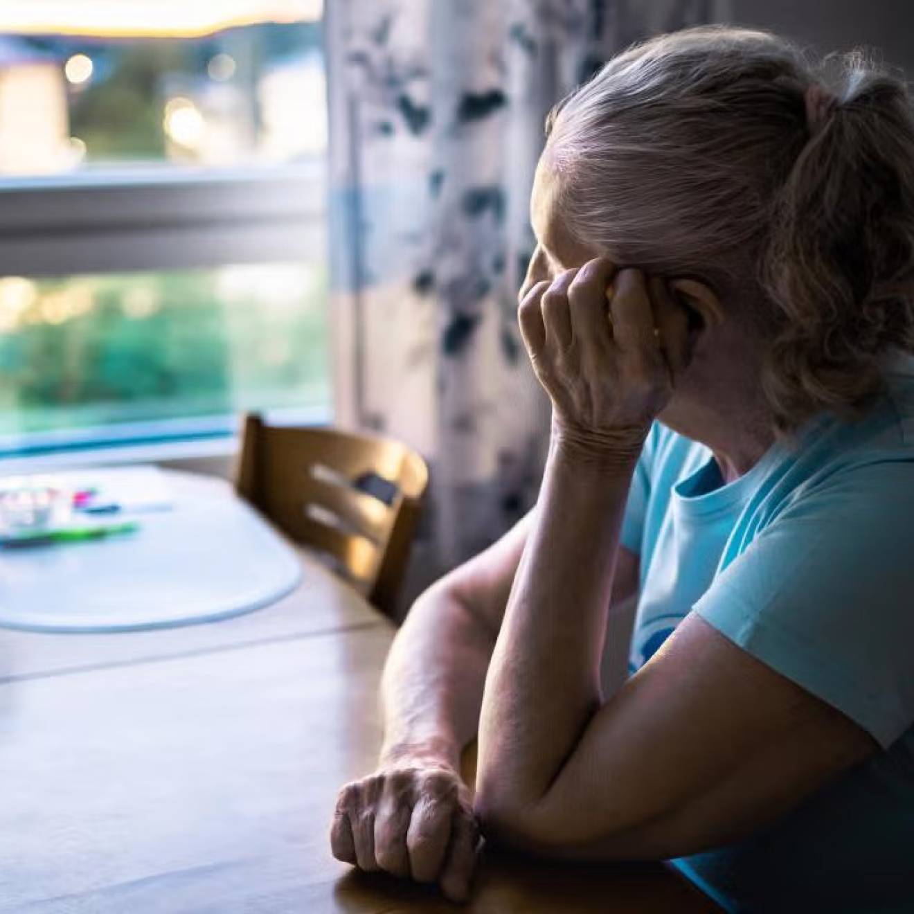 Older woman in blue shirt sitting at kitchen table, gazing pensively out window