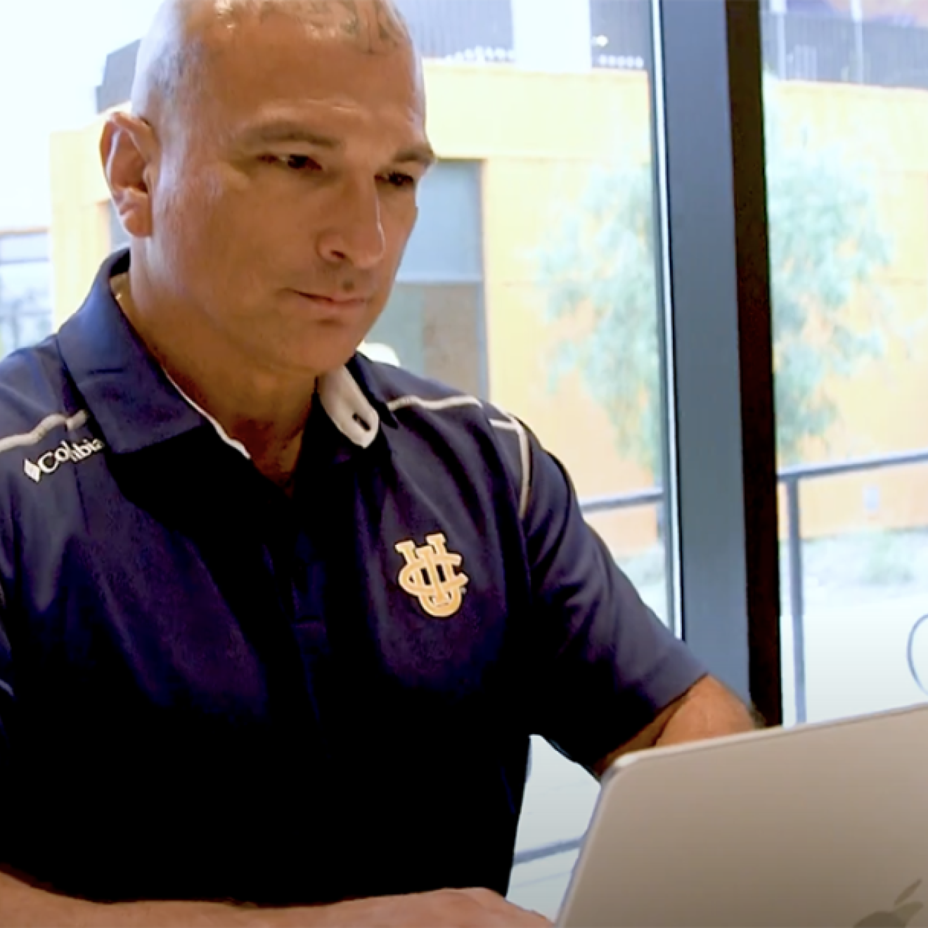 Patrick Acuña, wearing a blue UCI polo and with tattoos on his scalp, types on a laptop.