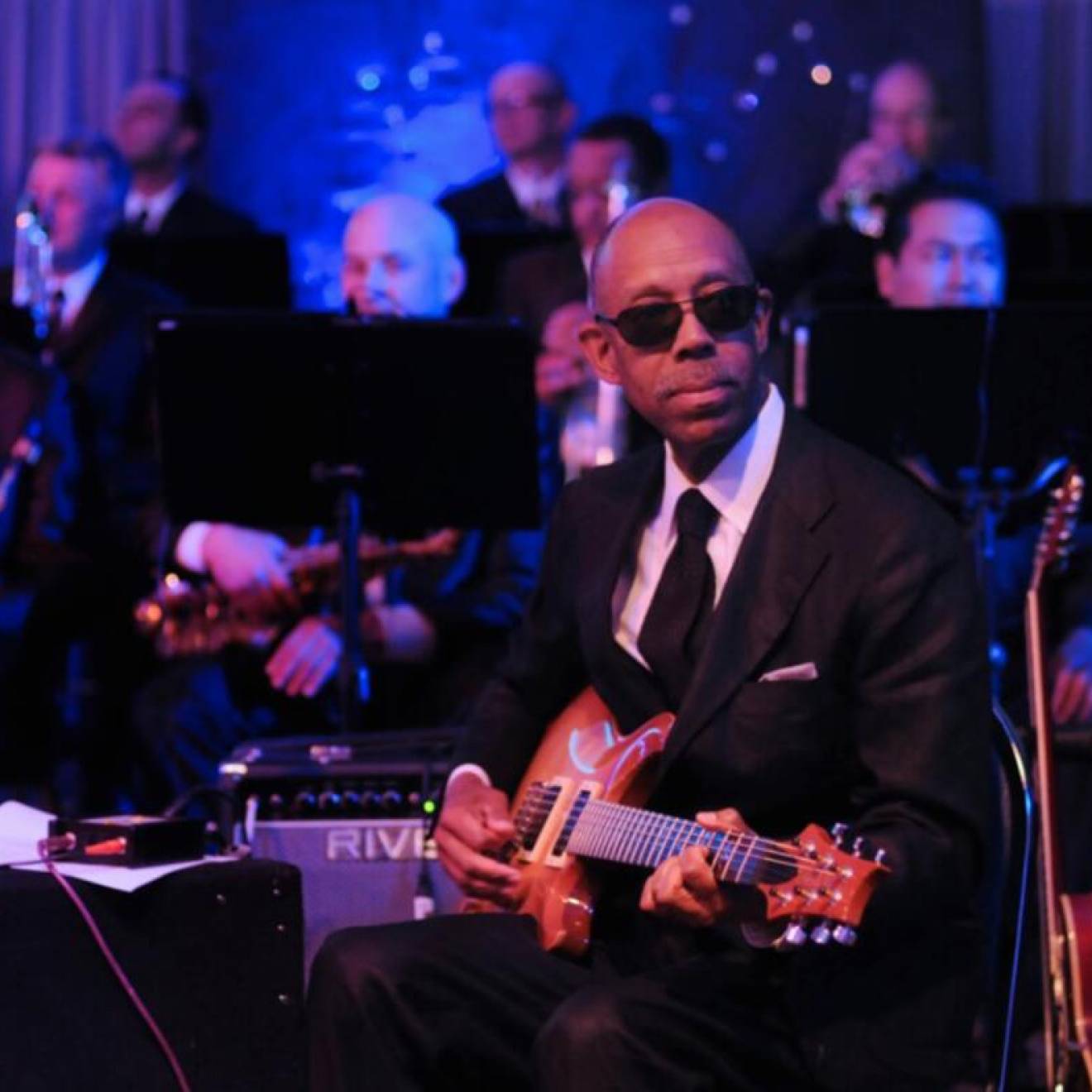 President Drake plays guitar on stage, wearing a black suit and sunglasses