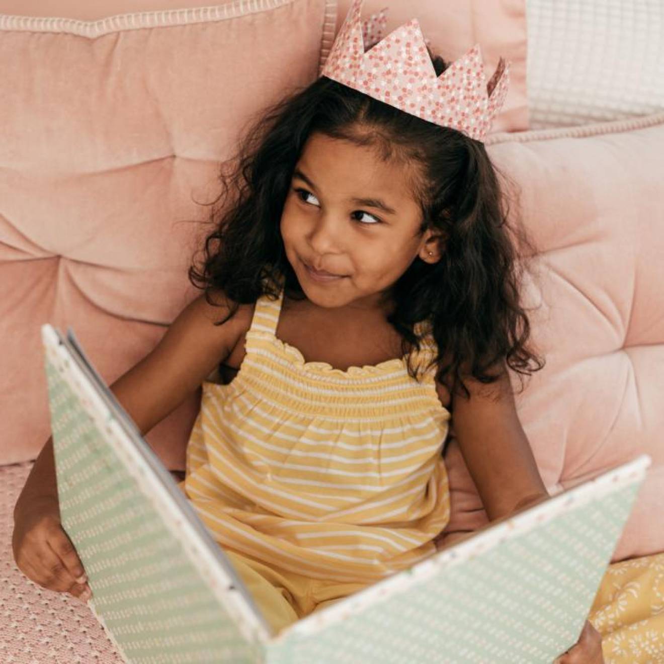 A young South Asian girl reads a book while wearing a crown and smiles