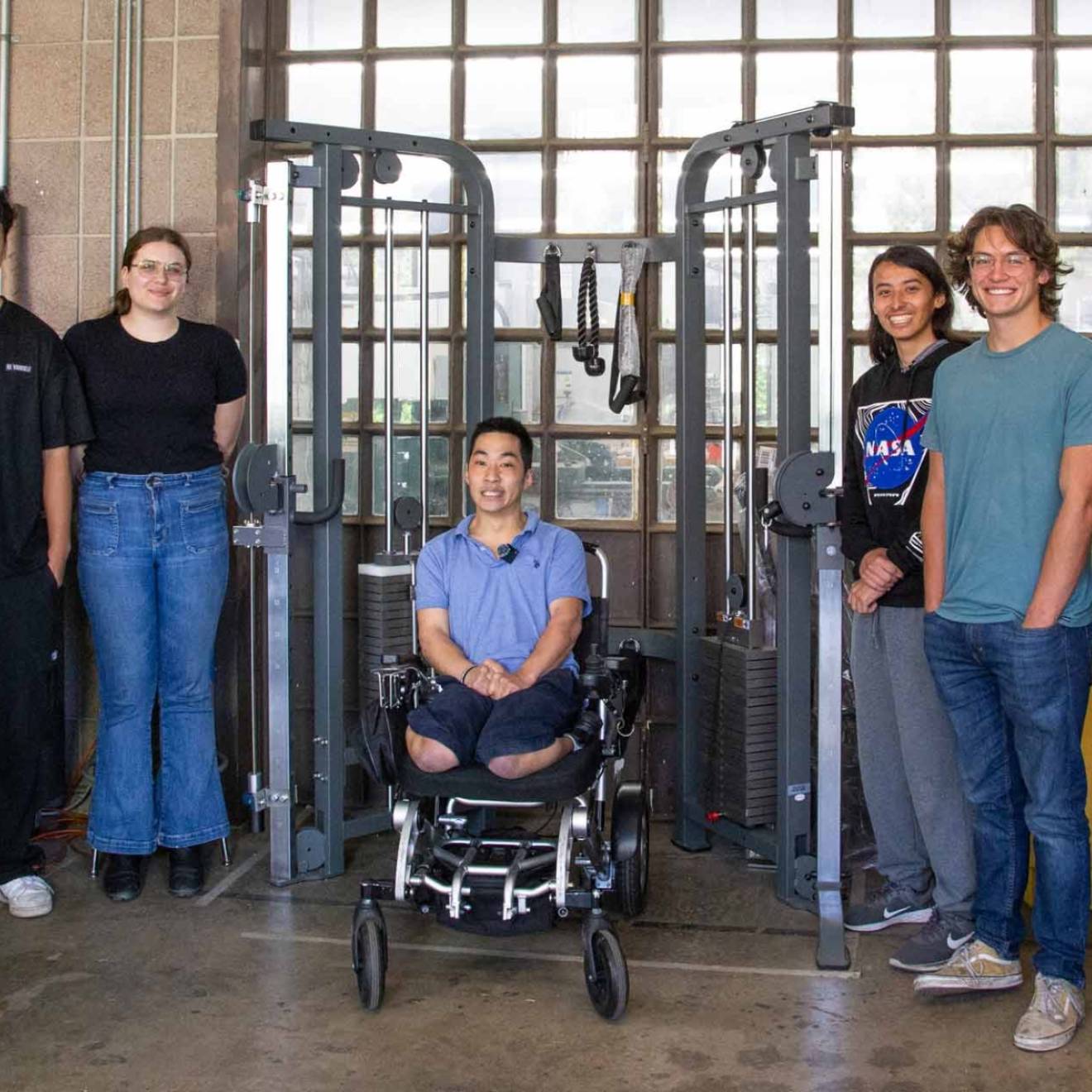 A team of students and a professor stands around a young man in a wheelchair underneath a workout machine