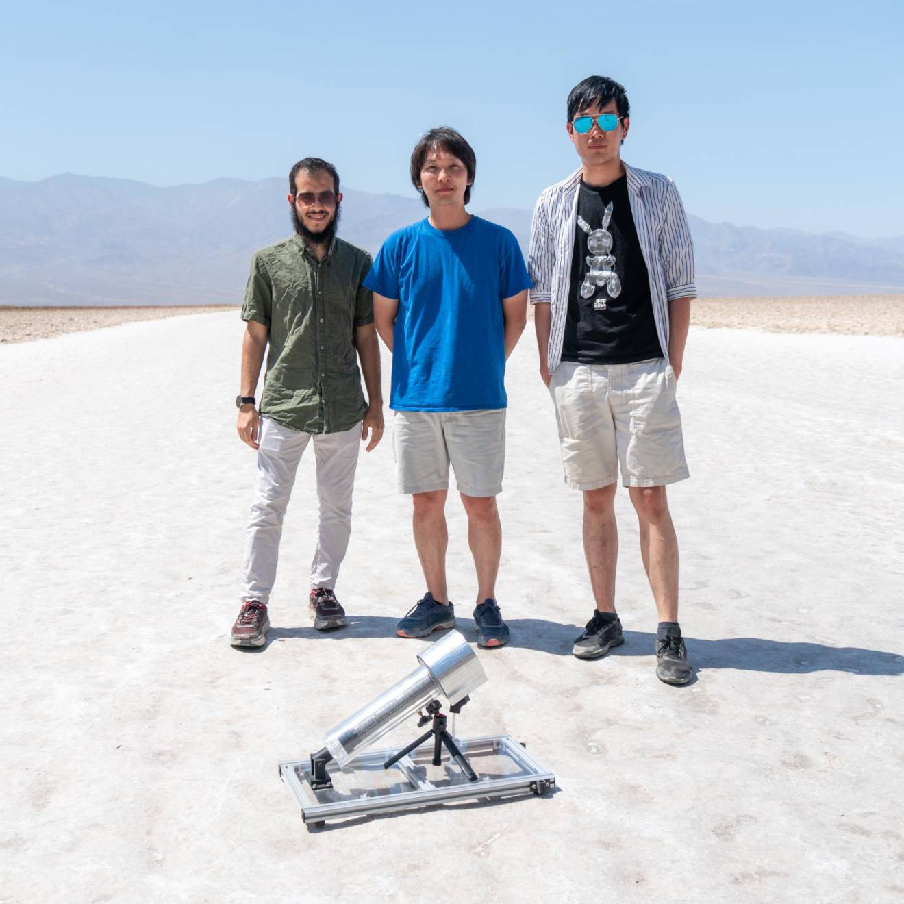 Three male students stand in white desert sands, behind a water harvester that looks like a microphone