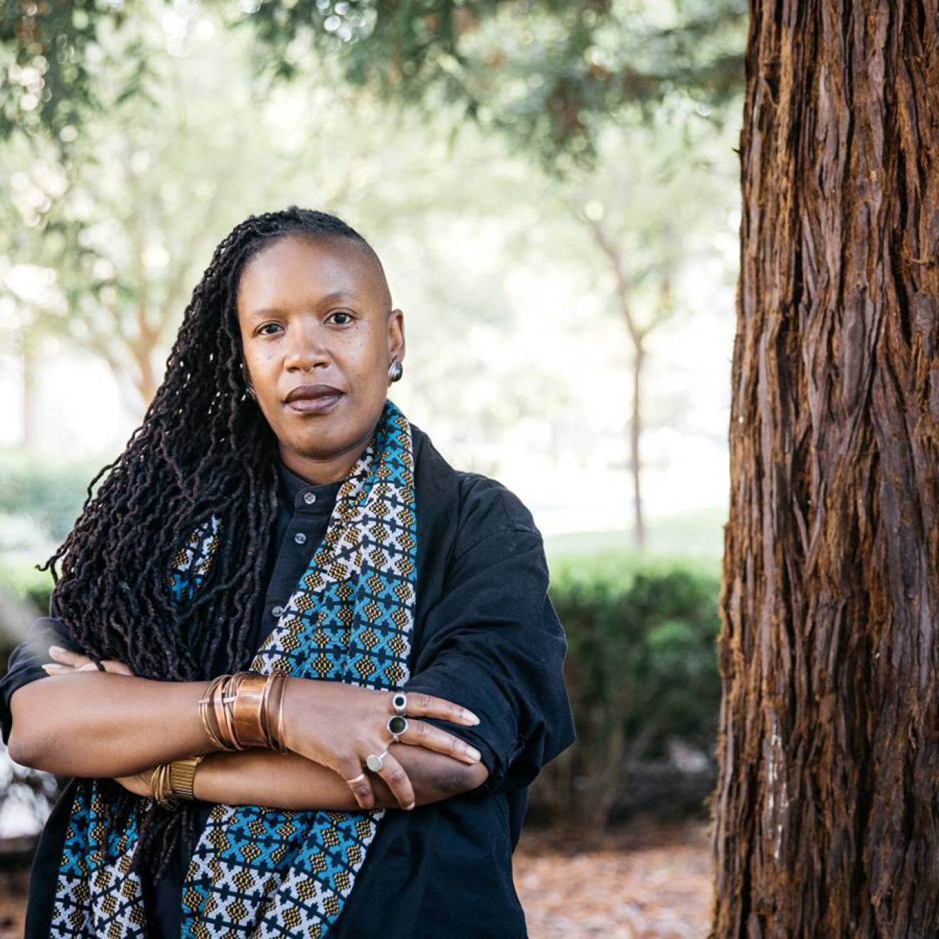 E. Tendayi Achiume, African American woman, poses outside, arms crossed, next to a redwood