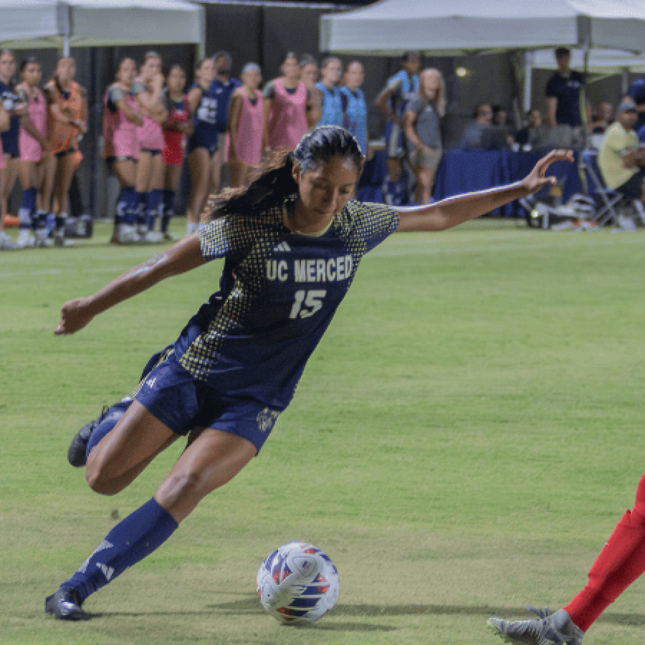 A woman in a blue UC Merced uniform prepares to kick the ball as a woman in a red uniform guards her