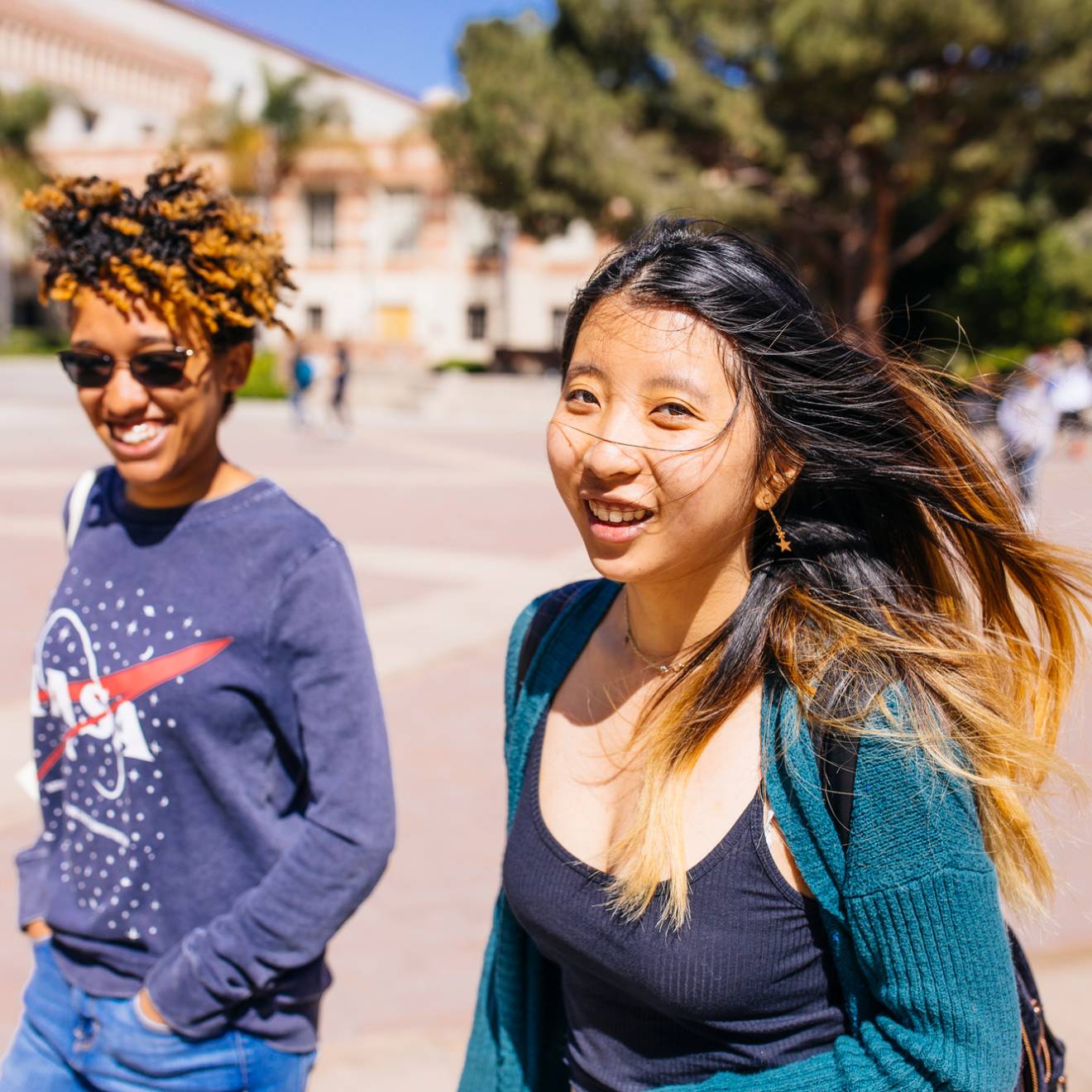 A young Black woman with a NASA sweatshirt and a young Asian woman with a green cardigan walk along a campus smiling