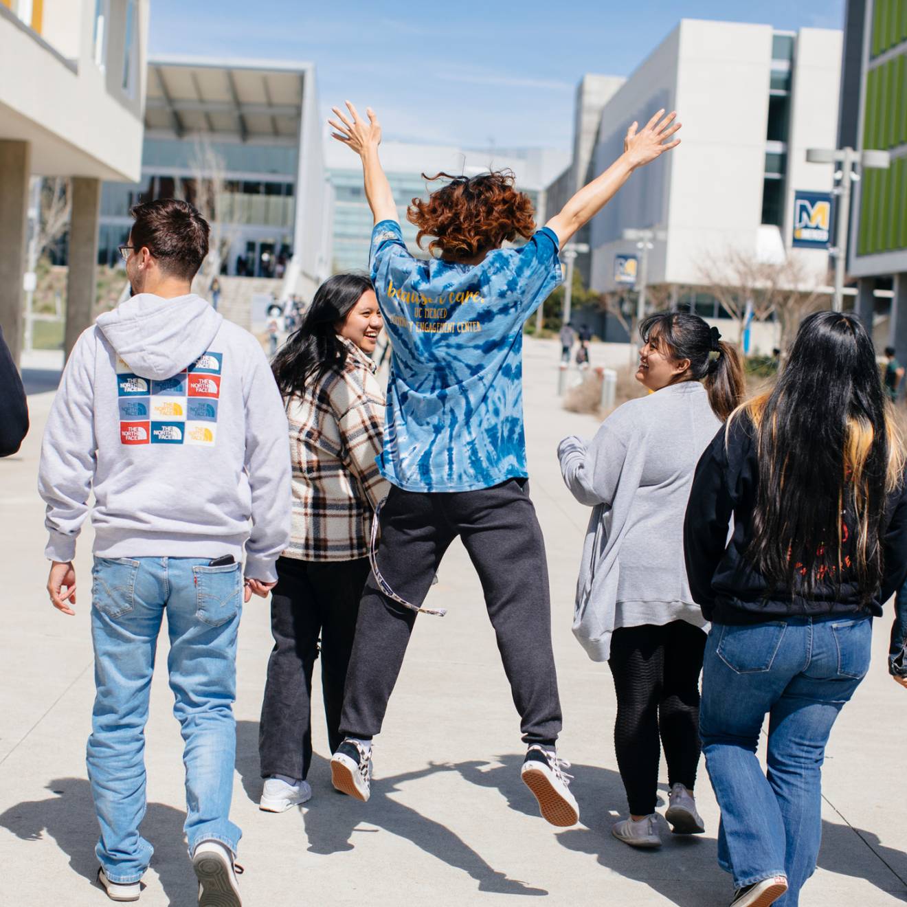 Viewed from behind, a diverse group of students walks along the UC Merced campus, the center student jumping exuberantly