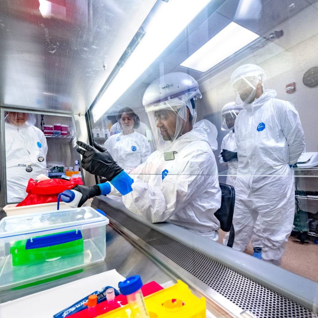 A group of students in PPE watch someone pour something behind protective glass