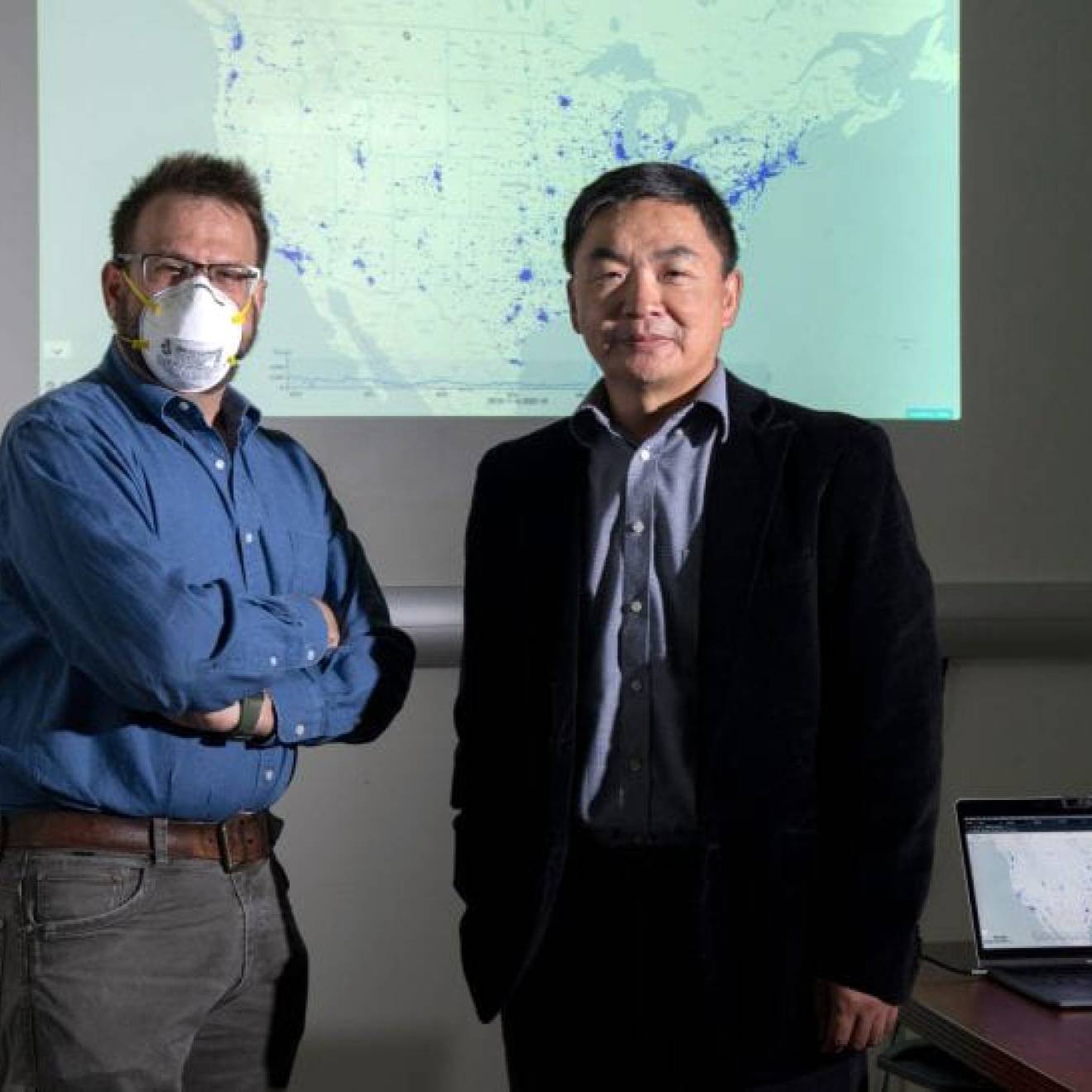 Two men standing together with a projector screen behind them, one wearing a face mask