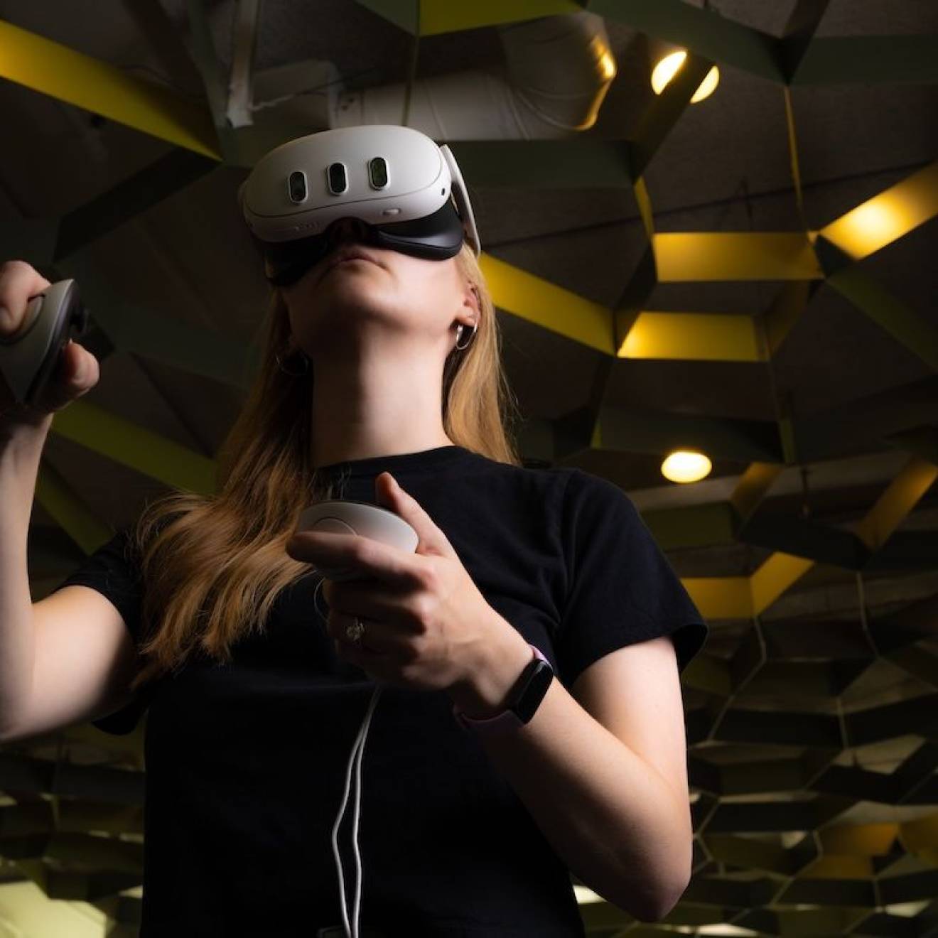 A student with a virtual reality headset and two controllers stands in a room with a honeycomb-esque roof