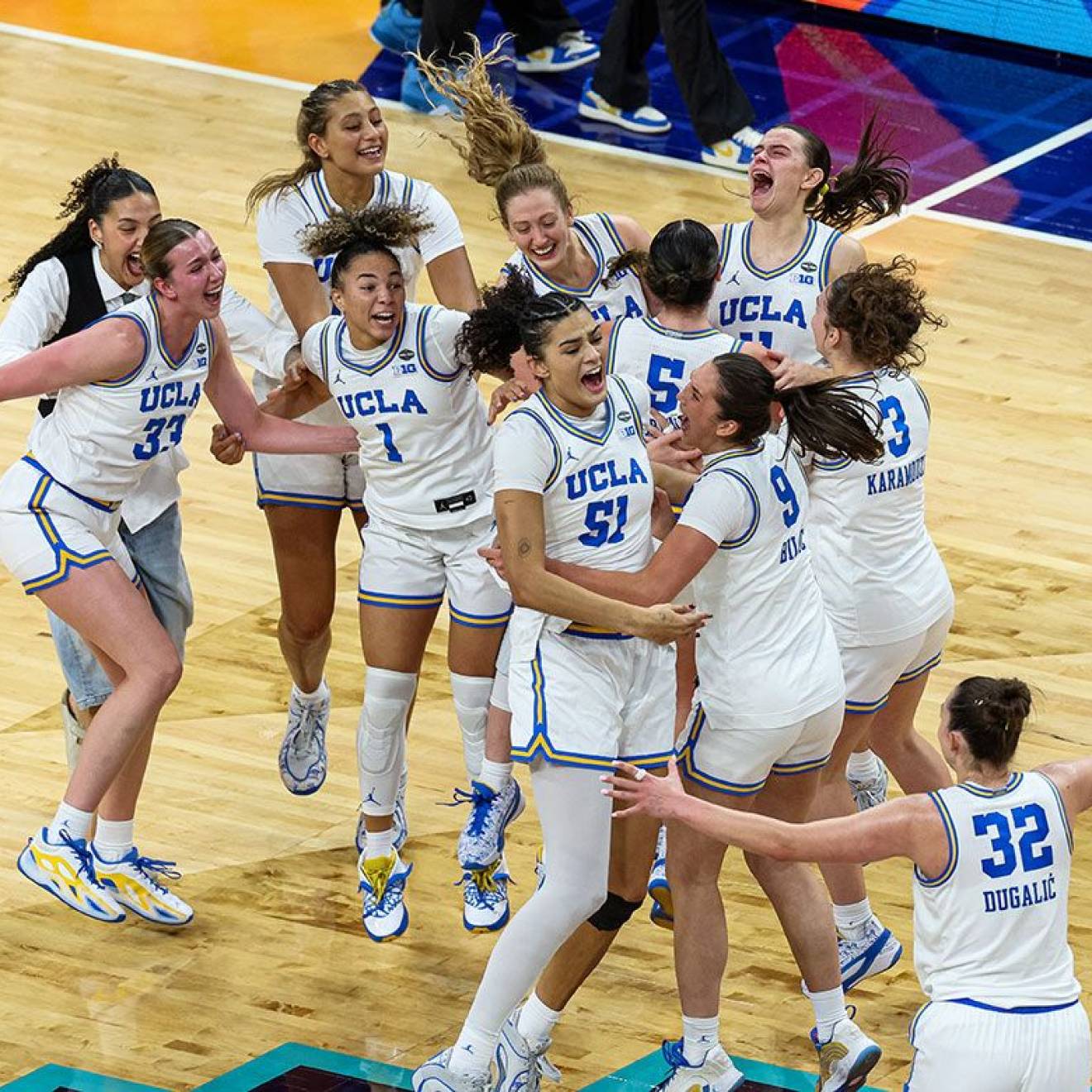 10 UCLA women's basketball players in jerseys and a woman in plain clothes jump on the court in celebration after winning the national championship