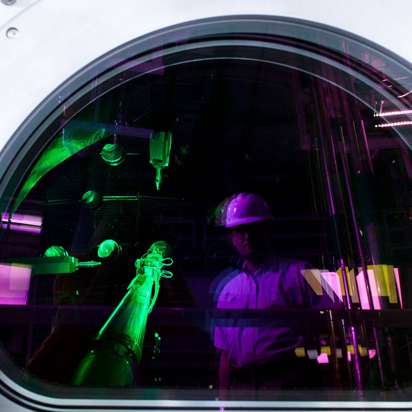 A person wearing a white hard hat is reflected in the circular viewing window of a large scientific instrument, while bright green laser beams and purple lighting illuminate the interior of a laboratory filled with optical equipment and metal piping.