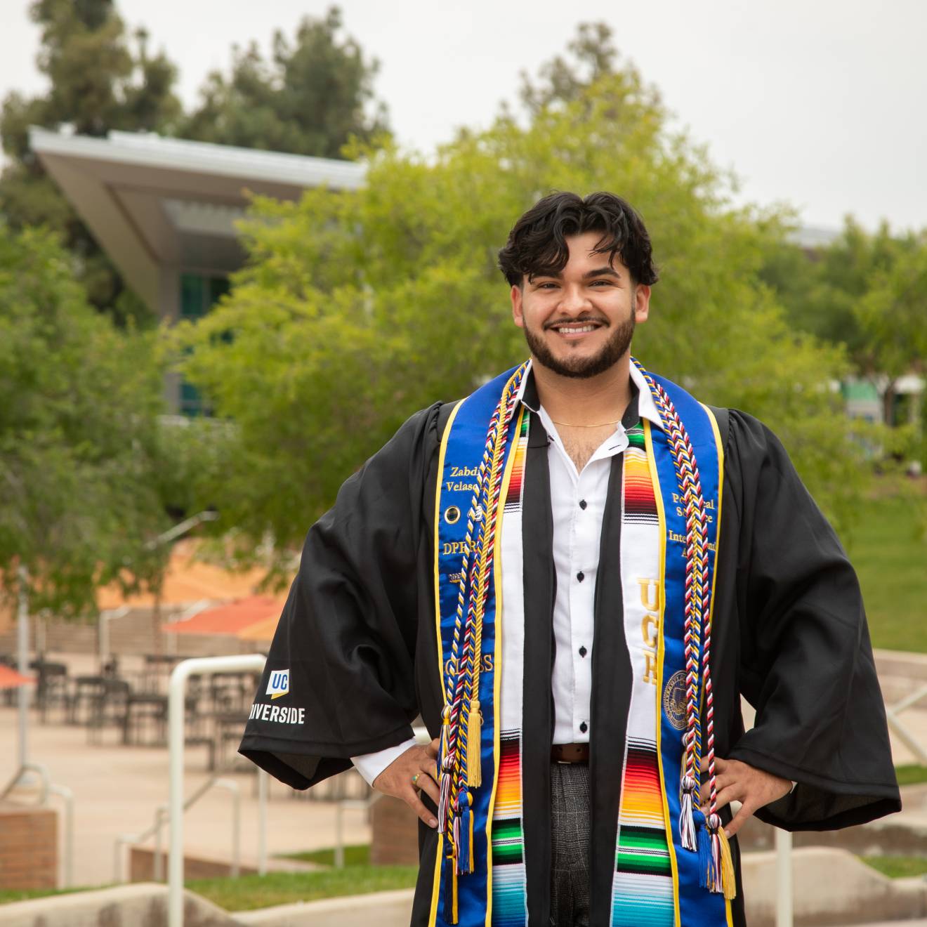 man with hands on hips wearing graduation gown and sashes on UC Riverside campus