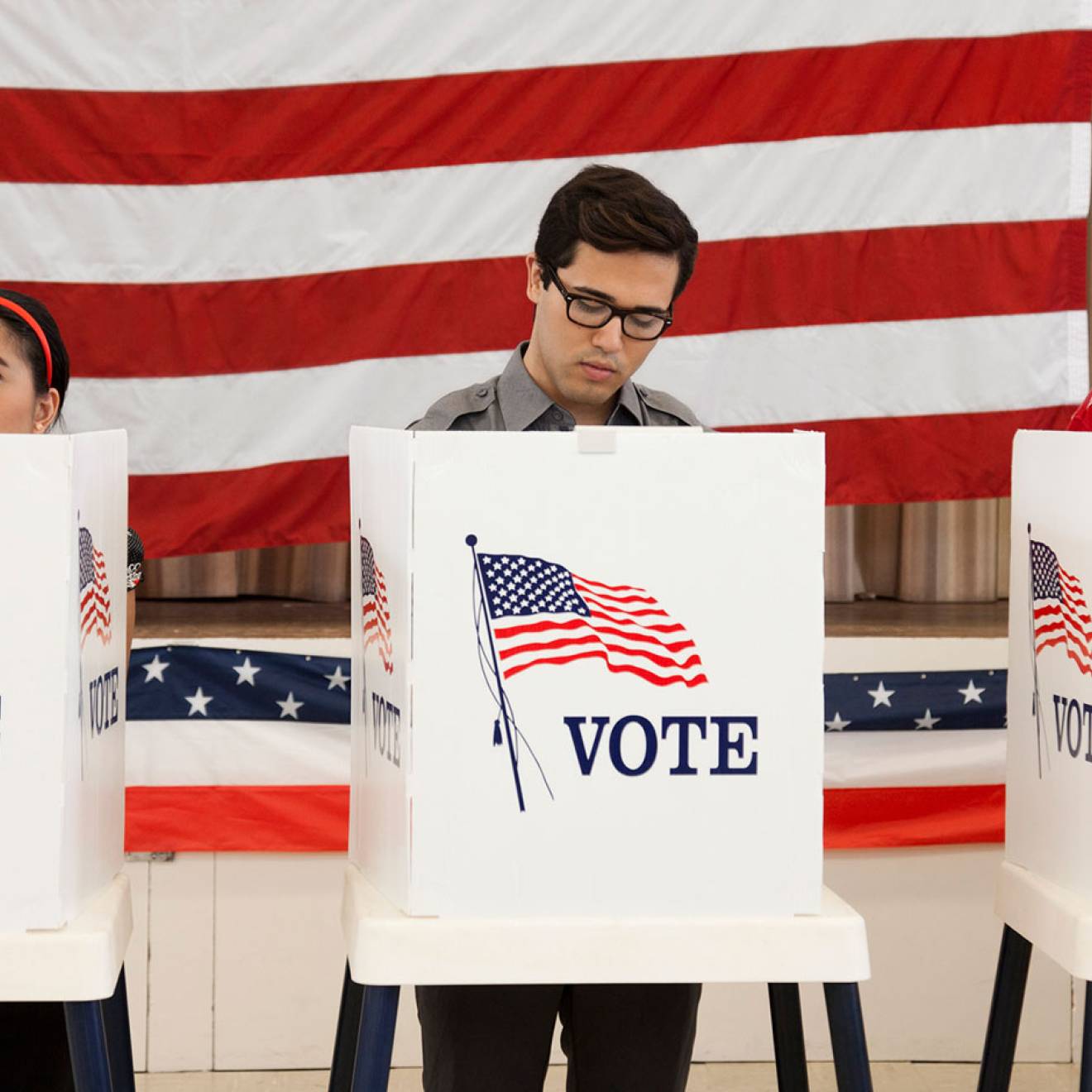 3 people stand at voting booths in front of a wall-sized American flag