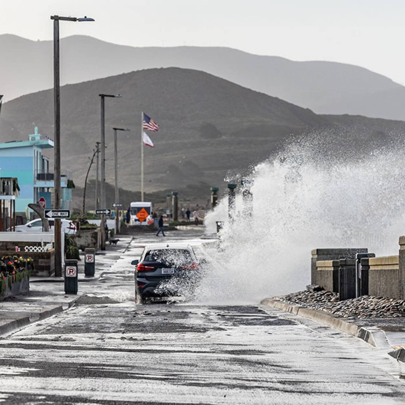 A wave crashes into a town street a blue car is driving on in Pacifica