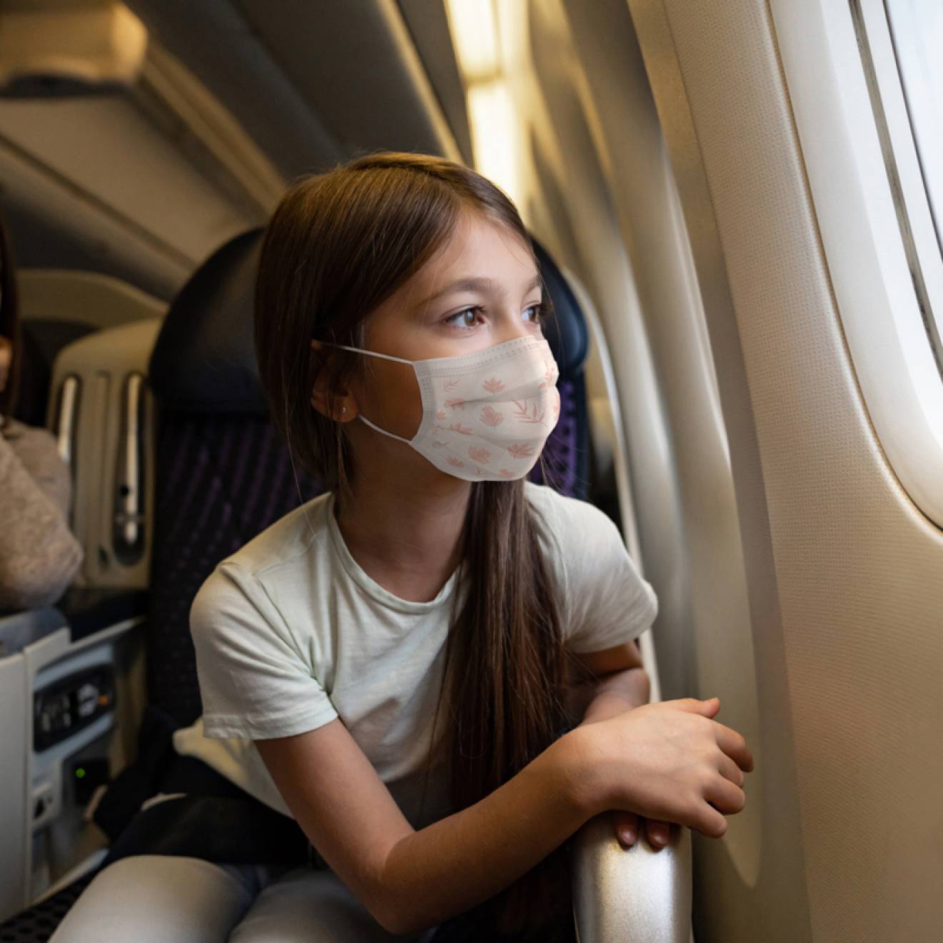 A young girl in a mask stares out an airplane window, mom in background