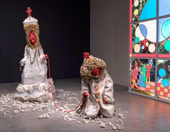 Sculptures in exhibit with white costumes and orange faces with a stained glass window in the background to the side