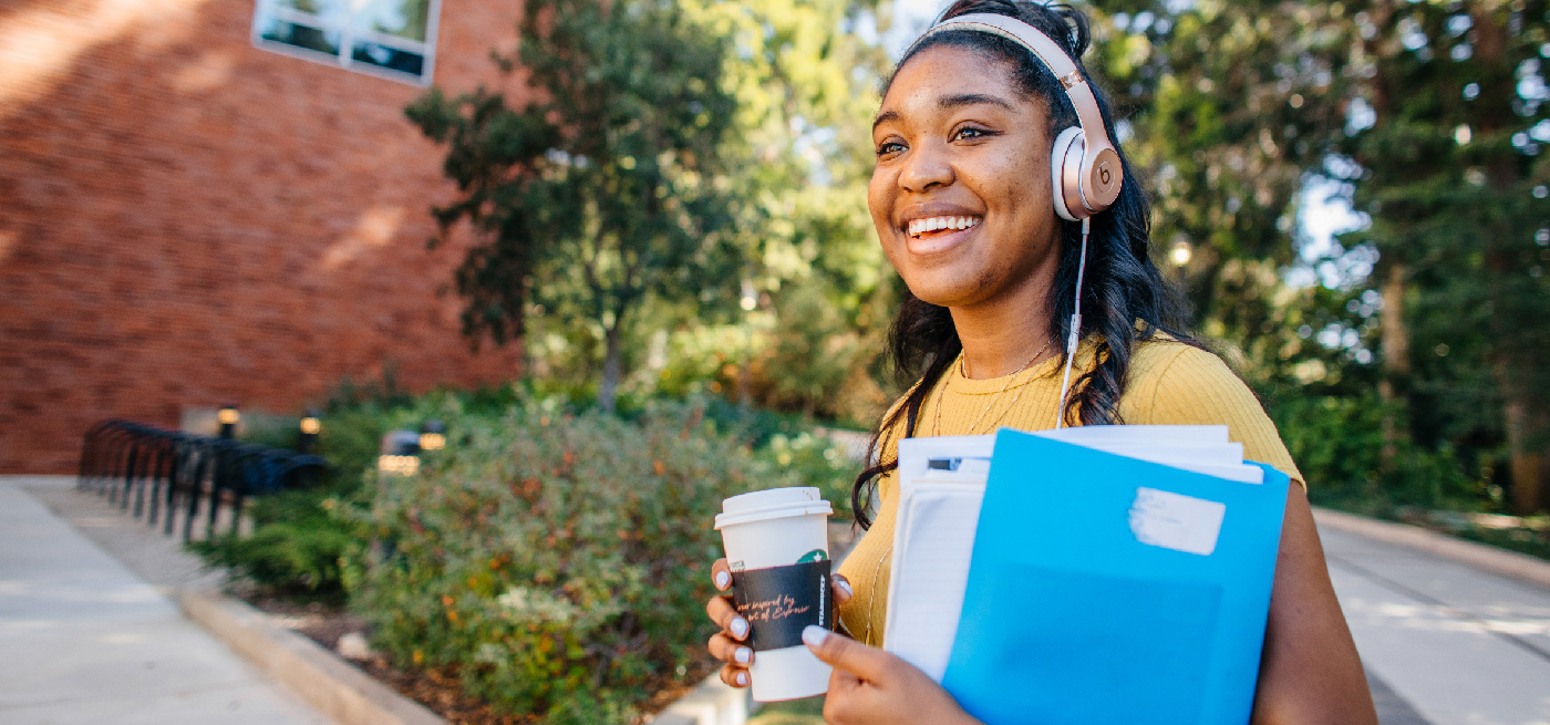 A student on campus, smiling, wearing headphones, holding a coffee and a folder of papers