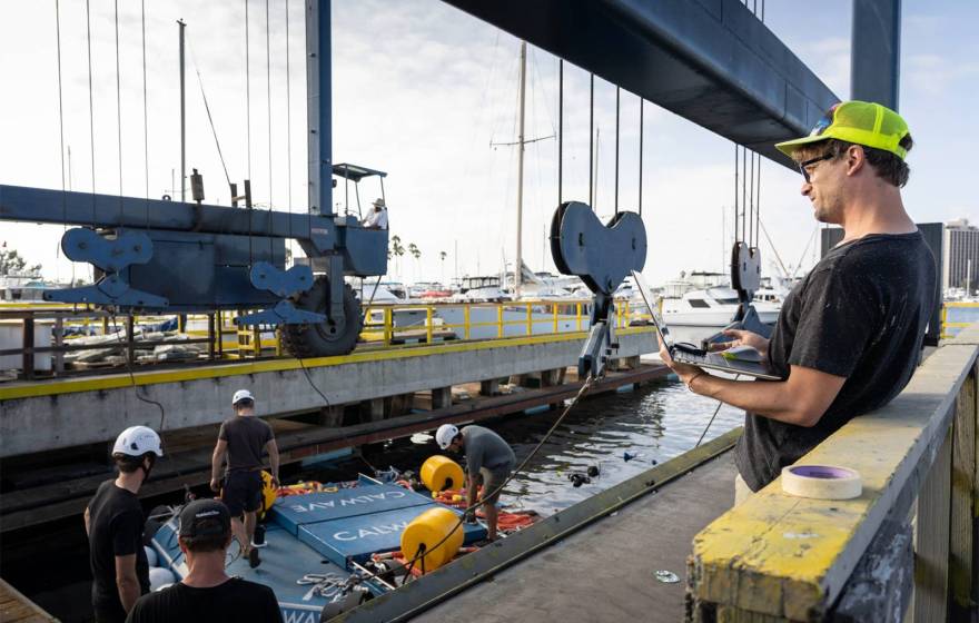People in hard hats work under a crane on a dock, with a blue, car-sized platform in the middle of the frame